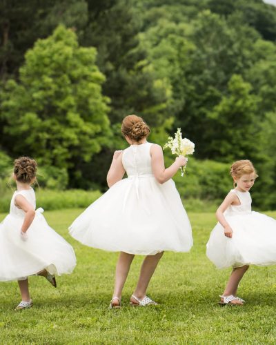 Three young flower girls with red hair pulled up in a bun wearing white dresses  are happy at an outdoor wedding on a sunny summer day, Minnesota, USA